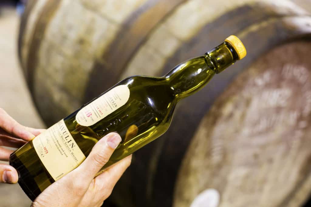 Someone holds a bottle of 16 years aged Lagavulin single malt whisky at the Lagavulin whisky distillery with old oak cask on the background.